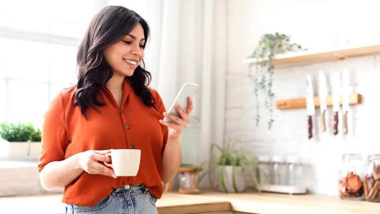 Woman holding a coffee and using her phone in a kitchen